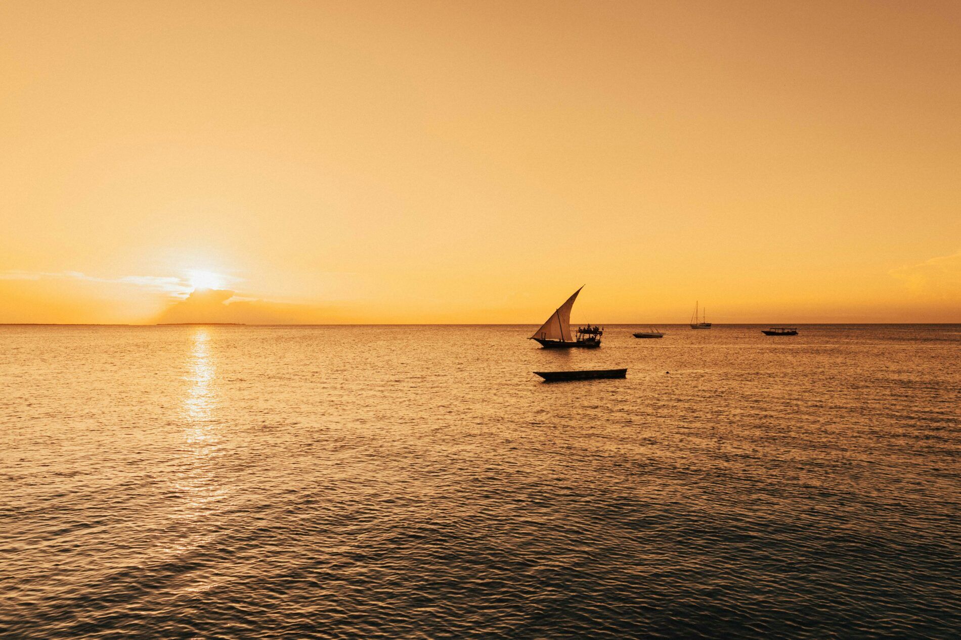 Dhow boats sale across calm ocean at sunset.