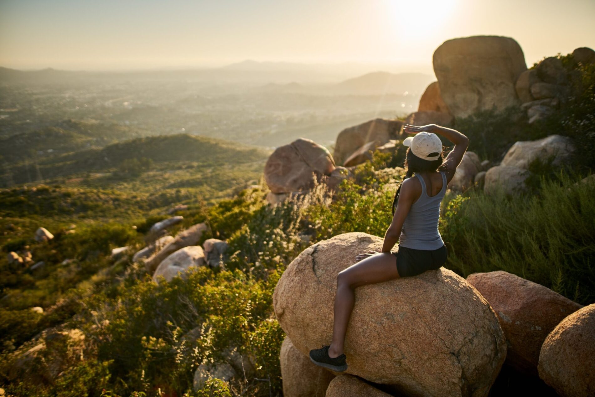Fit female hiker wearing cap sitting on rock at mountain top watching sunset