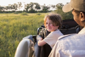 Young boy sits on guides lap while on safari vehicle and smiles at camera.