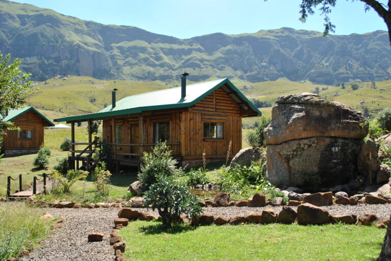 the outside of a log cabin with a boulder next to it with greenery and mountains in the background