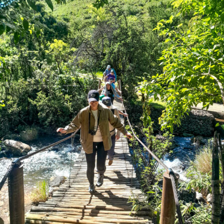 a group of tourists walking over a suspension bridge with water and greenery in the background