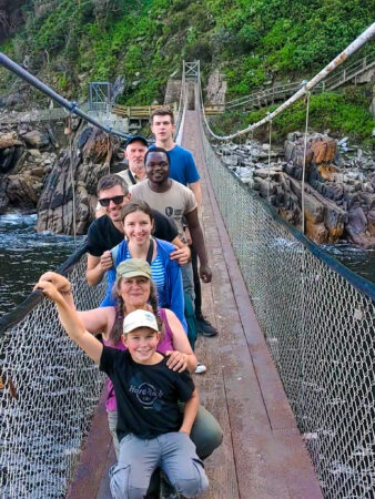 a group of tourists posing on a suspension bridge with water and greenery in the background