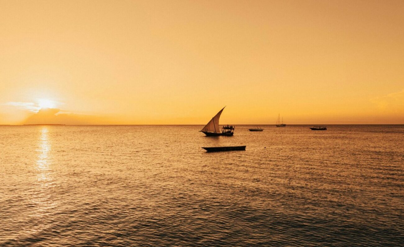 Dhow boats sale across calm ocean at sunset.